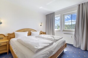 Ein ordentlich bezogenes Bett mit weißen Leinen, ein hölzernes Kopfende, ein Nachttisch mit Lampe und ein Fenster mit Vorhängen.