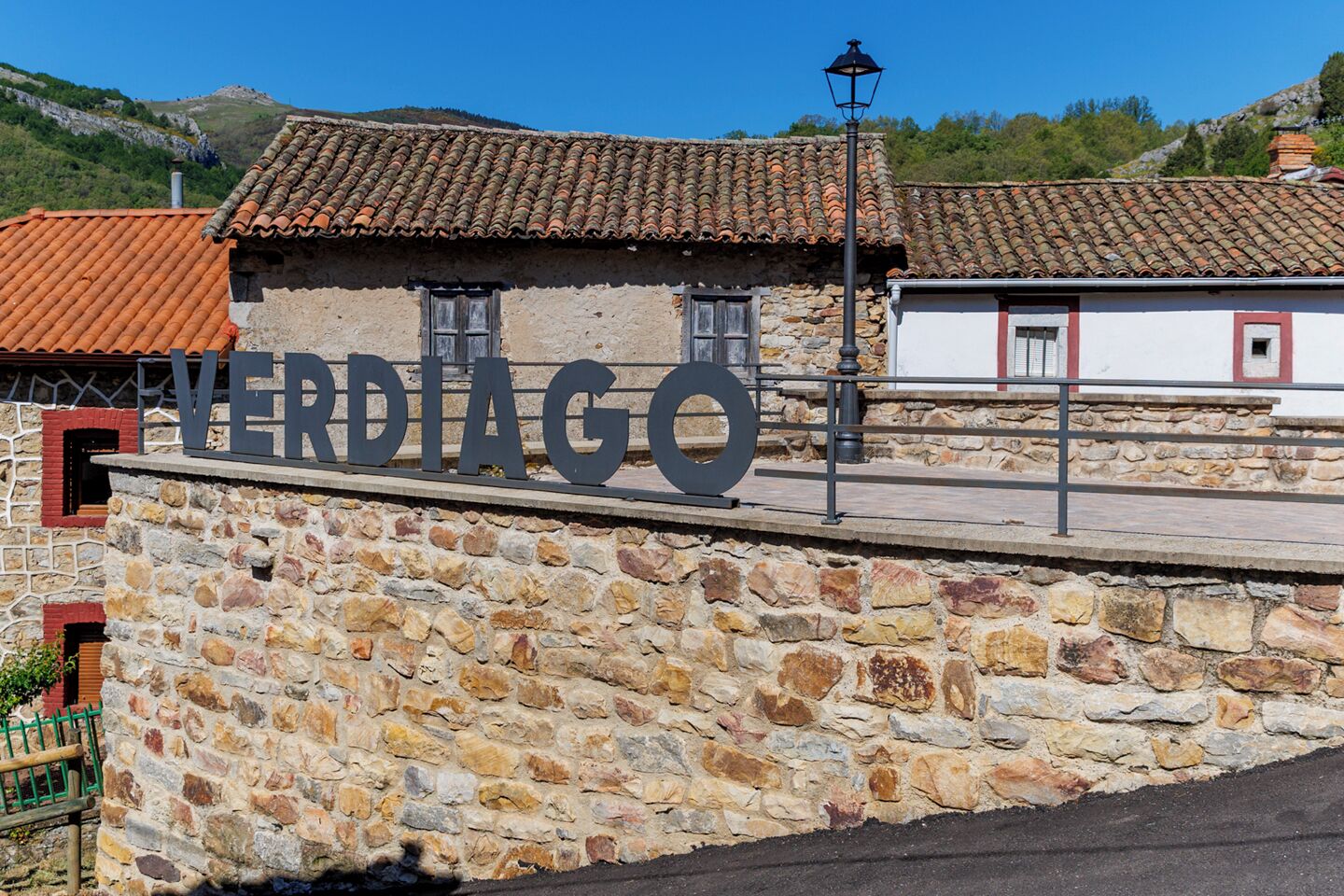 A stone wall with the name "VERDIGO" in large letters, a tiled roof, and a lamp post.