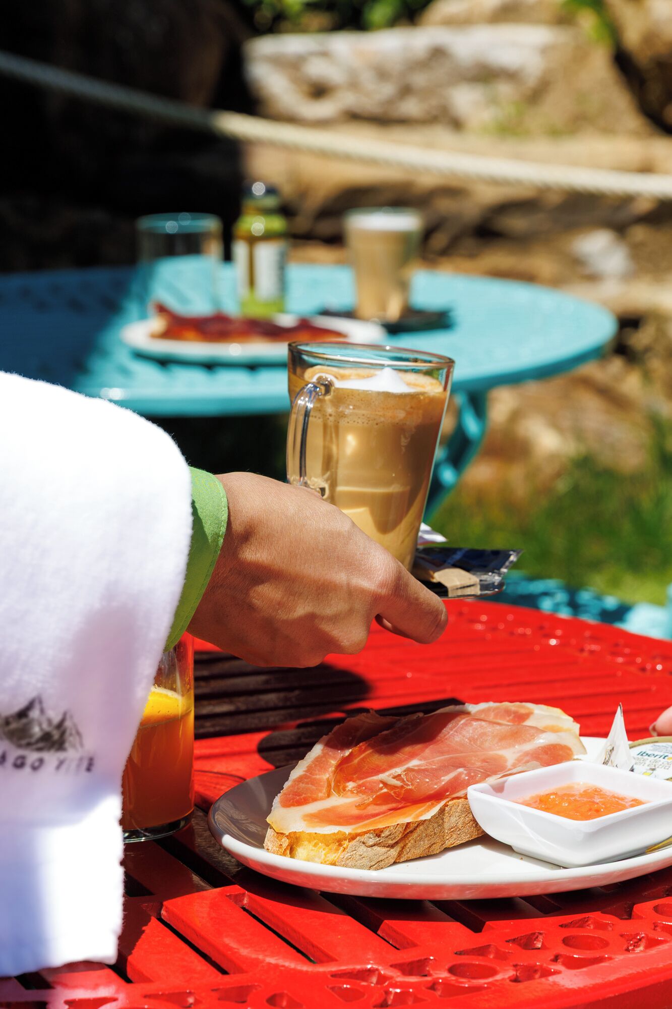 Una persona sosteniendo una taza de café, un plato con prosciutto y pan, y un recipiente pequeño con salsa, todo sobre una mesa roja al aire libre.