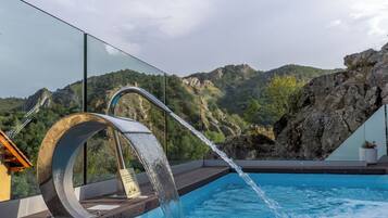 A modern outdoor pool with a waterfall feature, surrounded by a glass railing and overlooking a mountainous landscape.