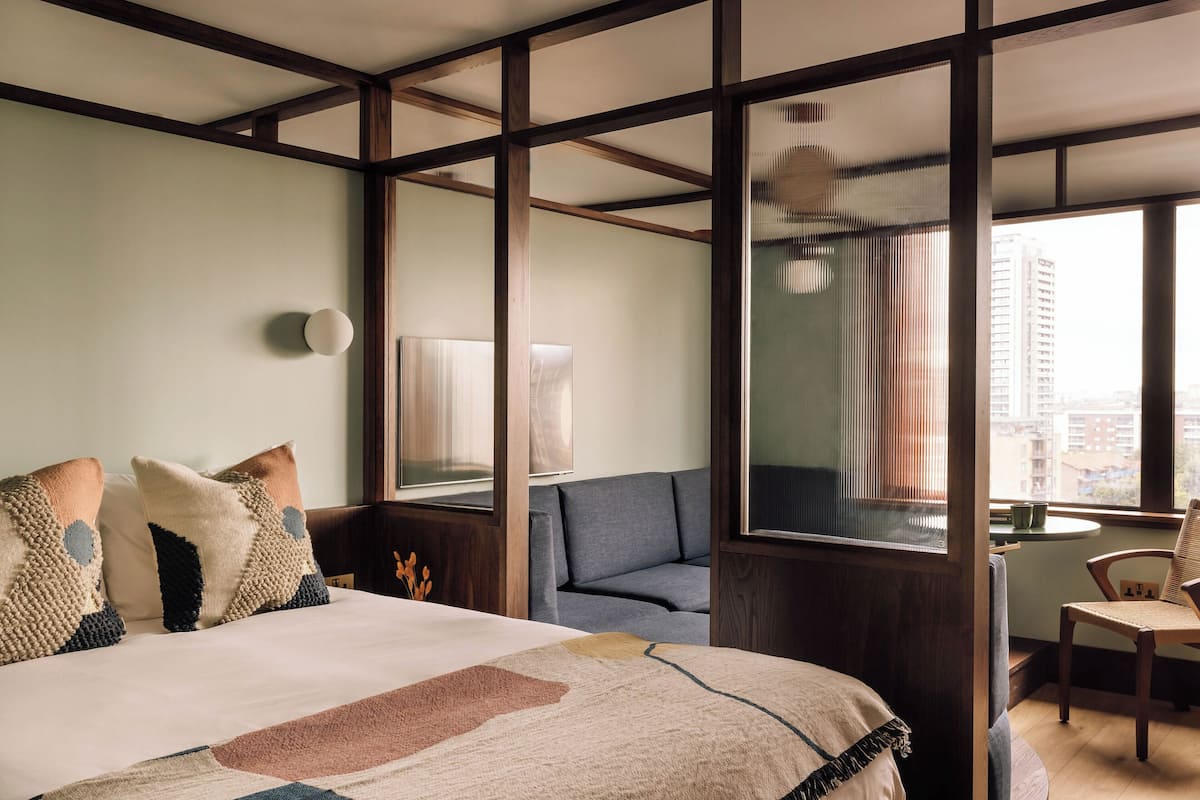 Ein Hotelzimmer mit einem großen Bett, einer Couch, einem Sessel und Blick auf die Stadt durch große Fenster.
