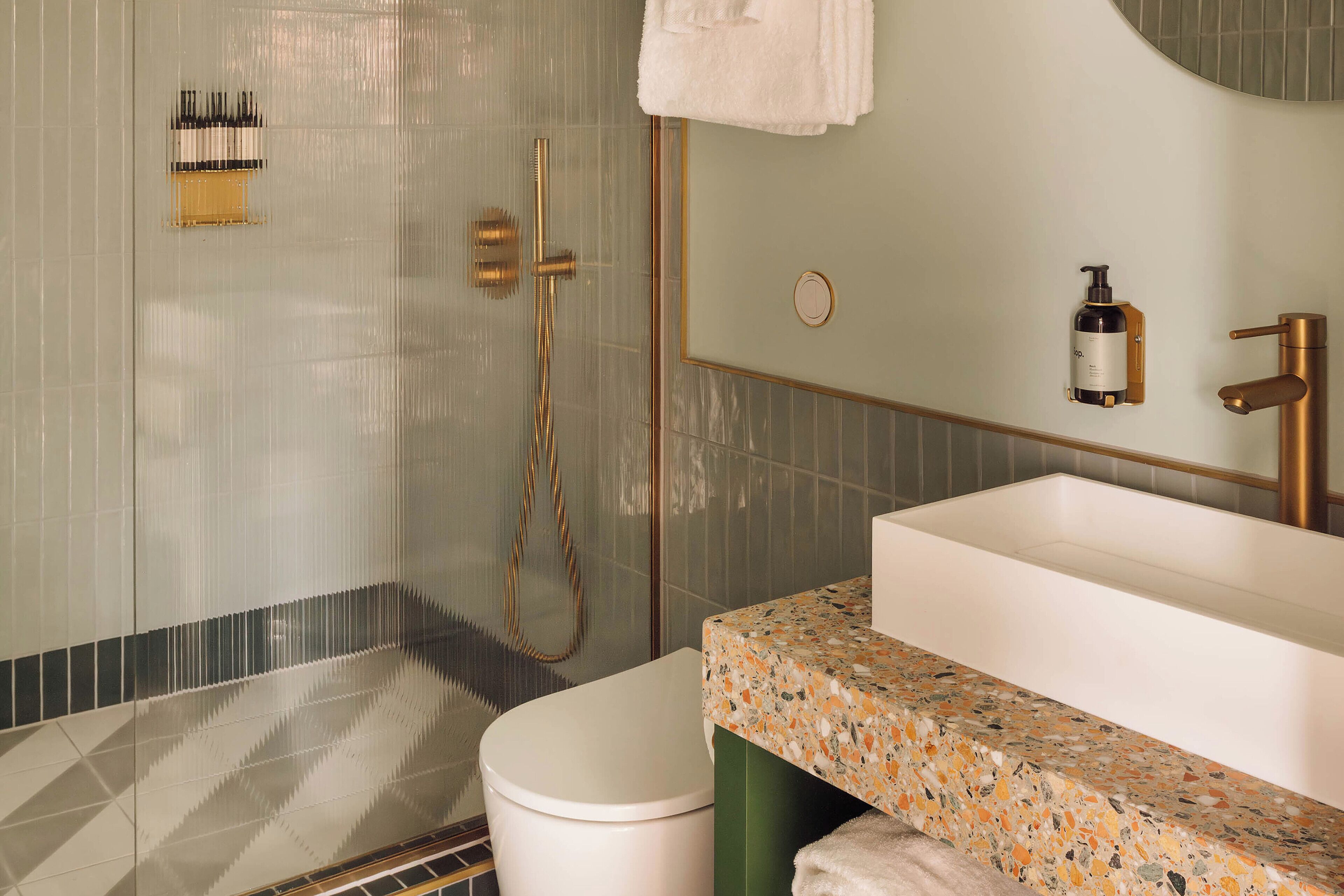 A bathroom with a white toilet, a shower with gold fixtures, and a sink with a speckled countertop.