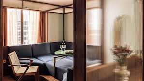 Un salon moderne avec un canapé, une chaise en bois, une petite table sur laquelle se trouvent une bouteille et des verres, et une grande fenêtre avec des rideaux.