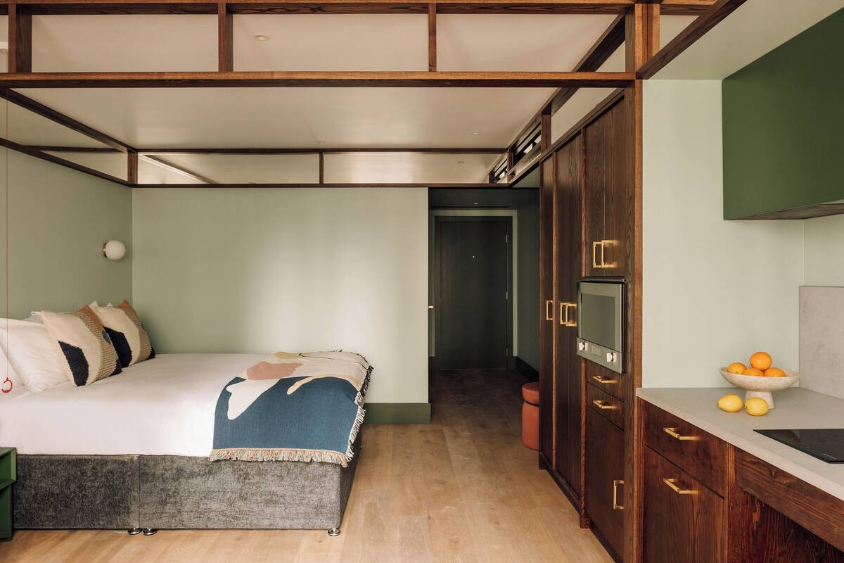 A bedroom with a bed, a bedside table, a wardrobe, and a small kitchen area with a sink and fruit bowl.