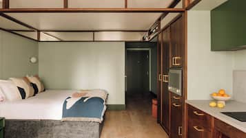 A bedroom with a bed, a bedside table, a wardrobe, and a small kitchen area with a sink and fruit bowl.