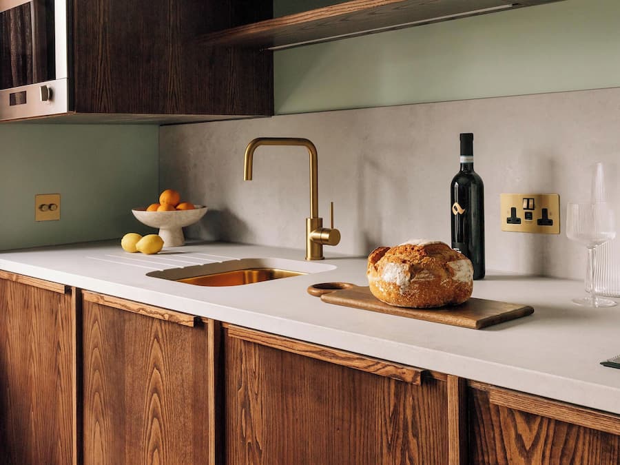Una cocina con un armario de madera, una encimera blanca, un grifo dorado, un bol de naranjas, una hogaza de pan, una botella de aceite de oliva y una copa de vino.