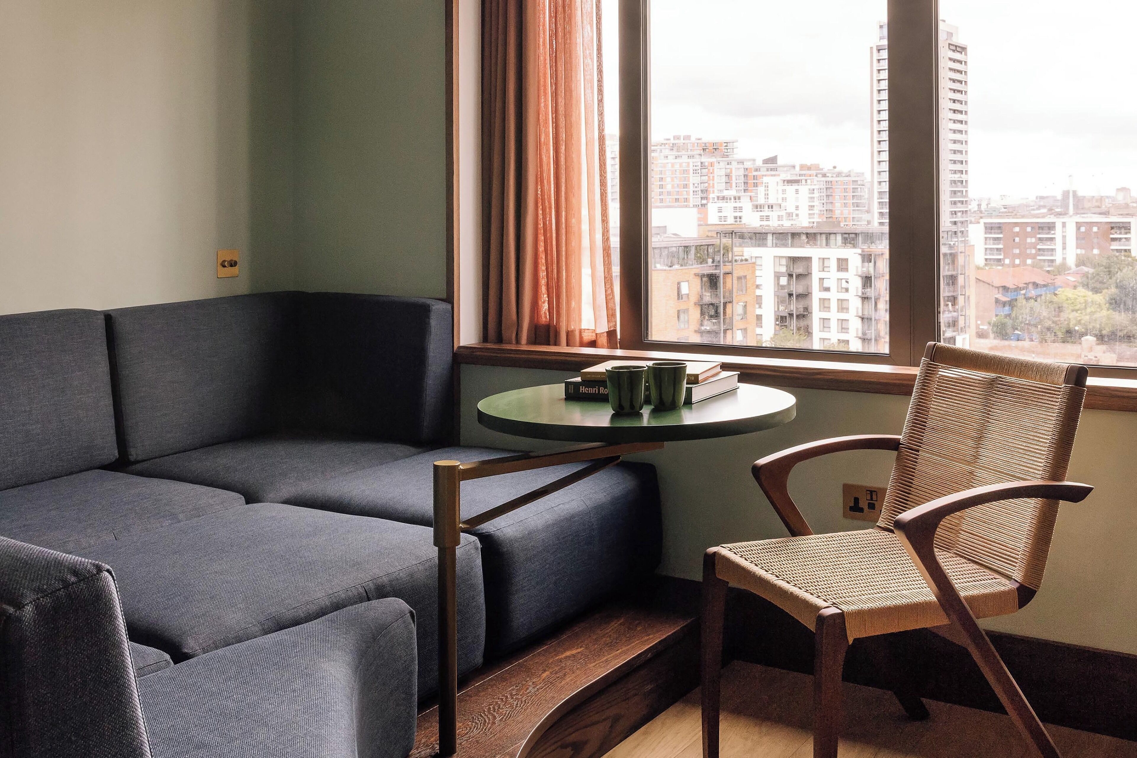 A modern living room with a grey sofa, a wooden chair, and a small table with books. A window with curtains provides a view of a cityscape.