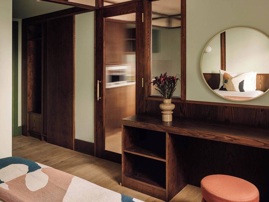 Una habitación con un armario de madera, un espejo redondo, una mesita con un jarrón y un otoman rosa.