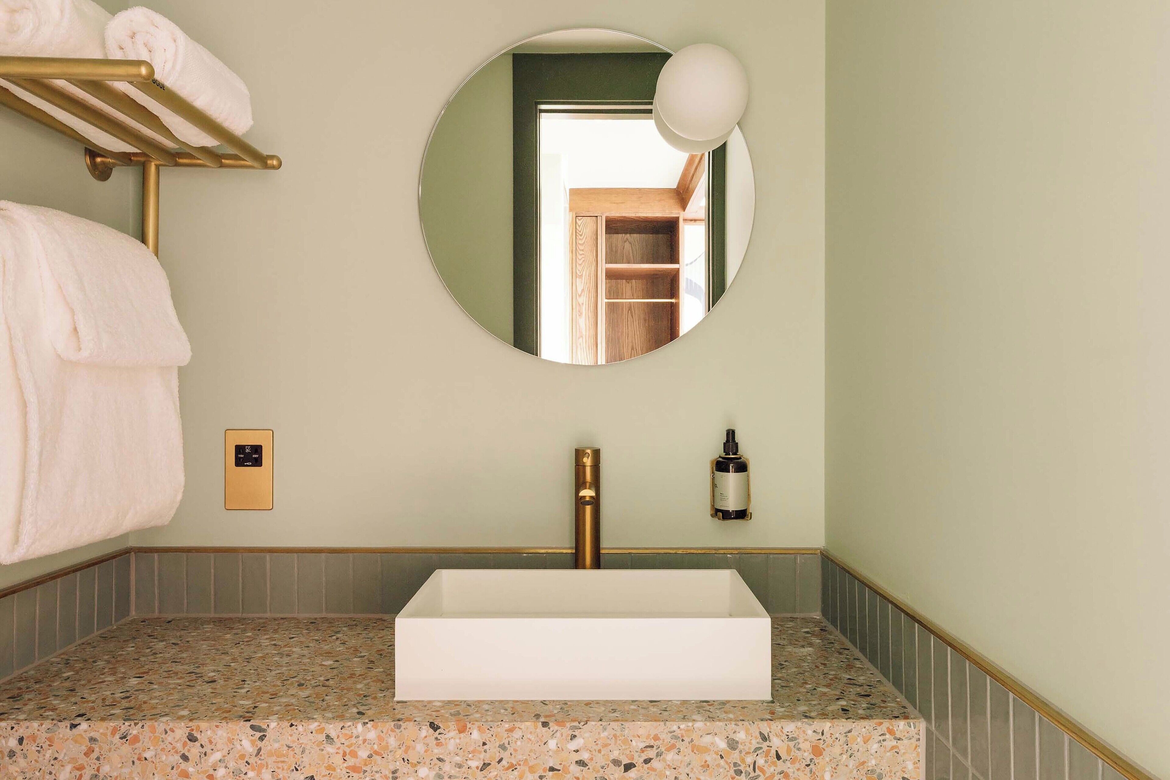 A bathroom with a rectangular sink, a gold towel rack, a round mirror, and a bottle of hand soap on the wall.