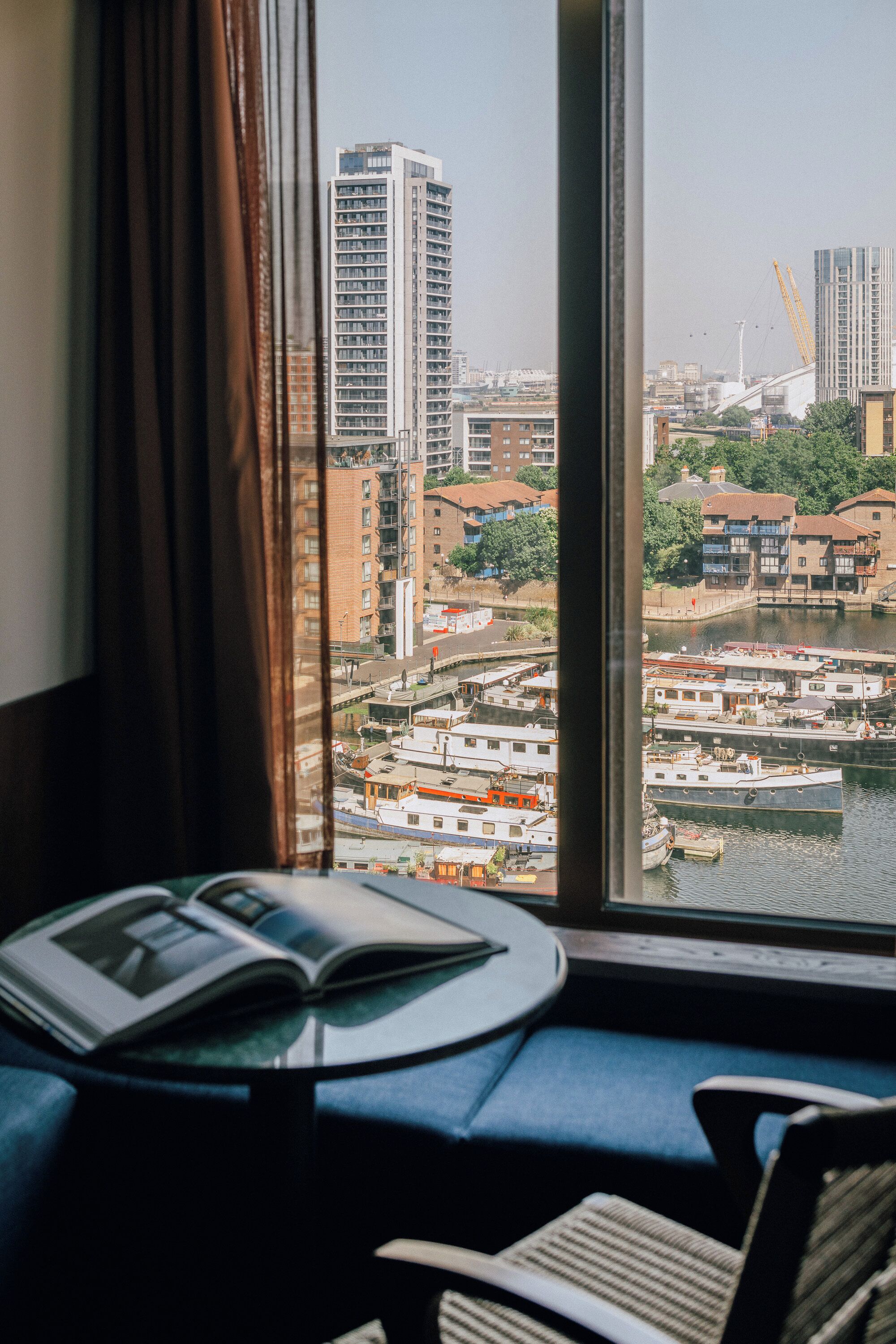 Habitación con vistas a un paisaje urbano a través de una ventana, que incluye una mesa con un libro y una silla.