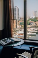 A room with a view of a cityscape through a window, featuring a table with a book and a chair.