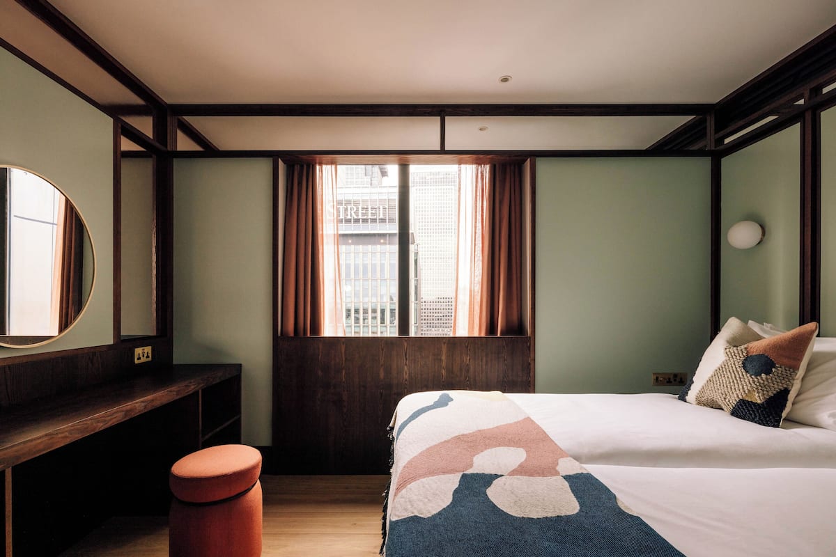 Ein Hotelzimmer mit einem großen Bett, einer hölzernen Bank, einem runden Spiegel und einem Fenster mit Vorhängen.