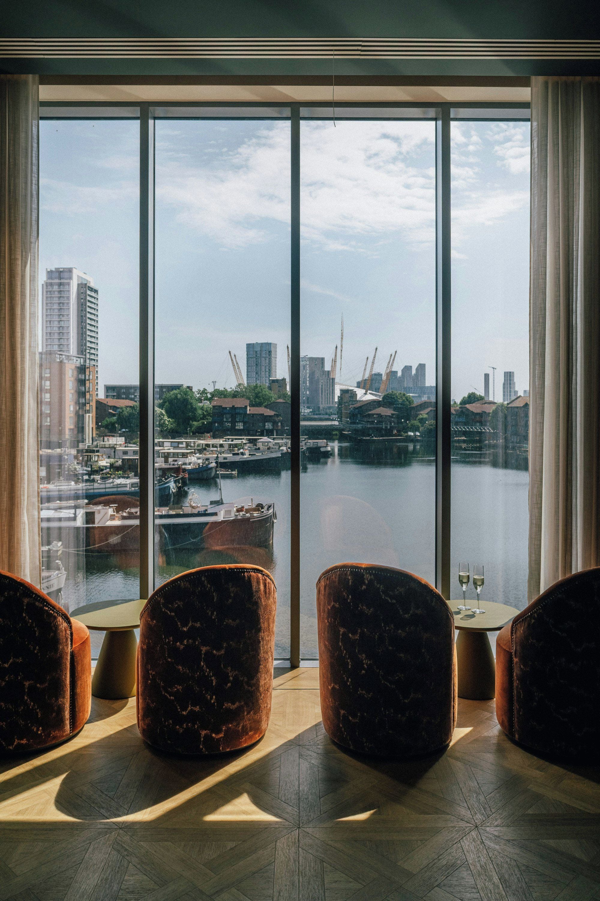 A room with a large window overlooking a marina and city skyline. There are three brown upholstered chairs and a small round table with two wine glasses.