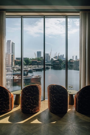 A room with a large window overlooking a marina and city skyline. There are three brown upholstered chairs and a small round table with two wine glasses. - Locke London Canary Wharf (London)