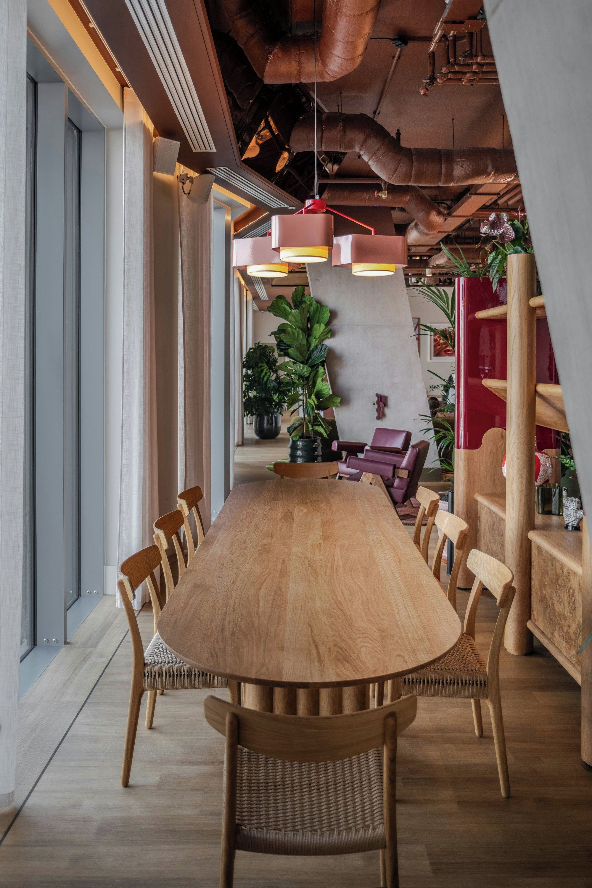 A modern dining area with a long wooden table surrounded by chairs, overhead industrial-style lighting, and potted plants.