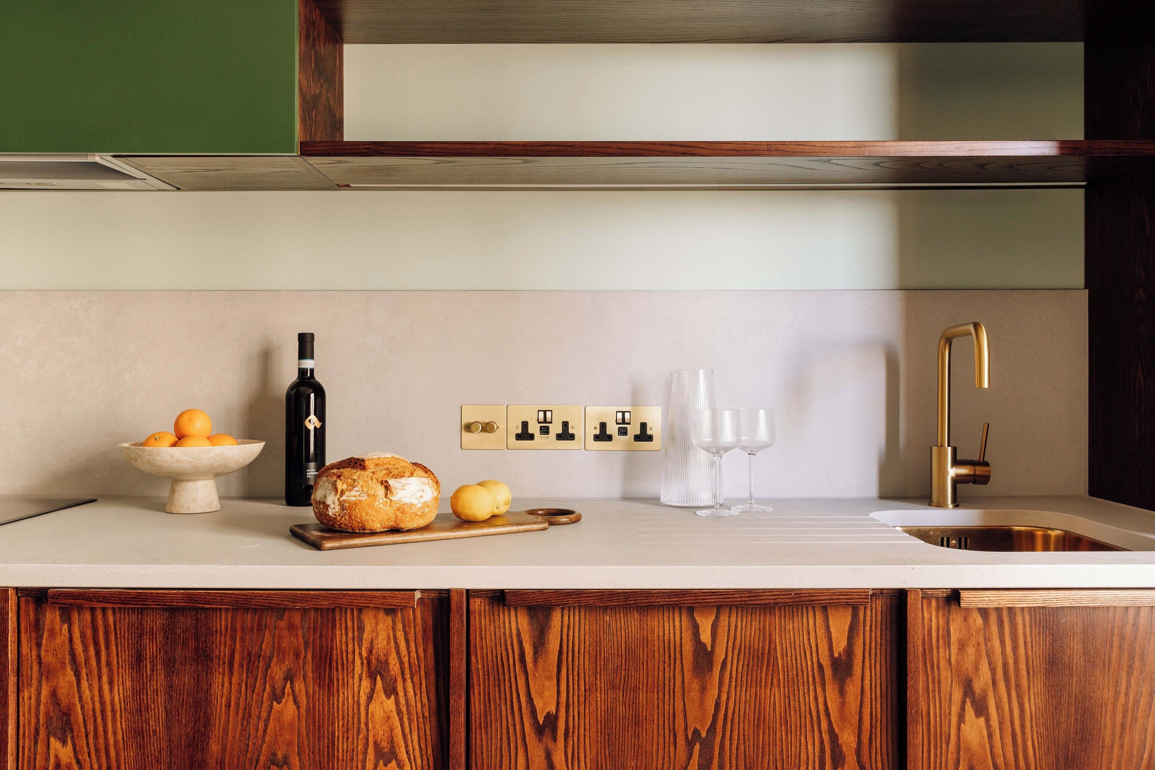 Una cocina con armarios de madera, una encimera, un bol de naranjas, una hogaza de pan, una botella de vino, una tabla de cortar y un lavabo con grifo.