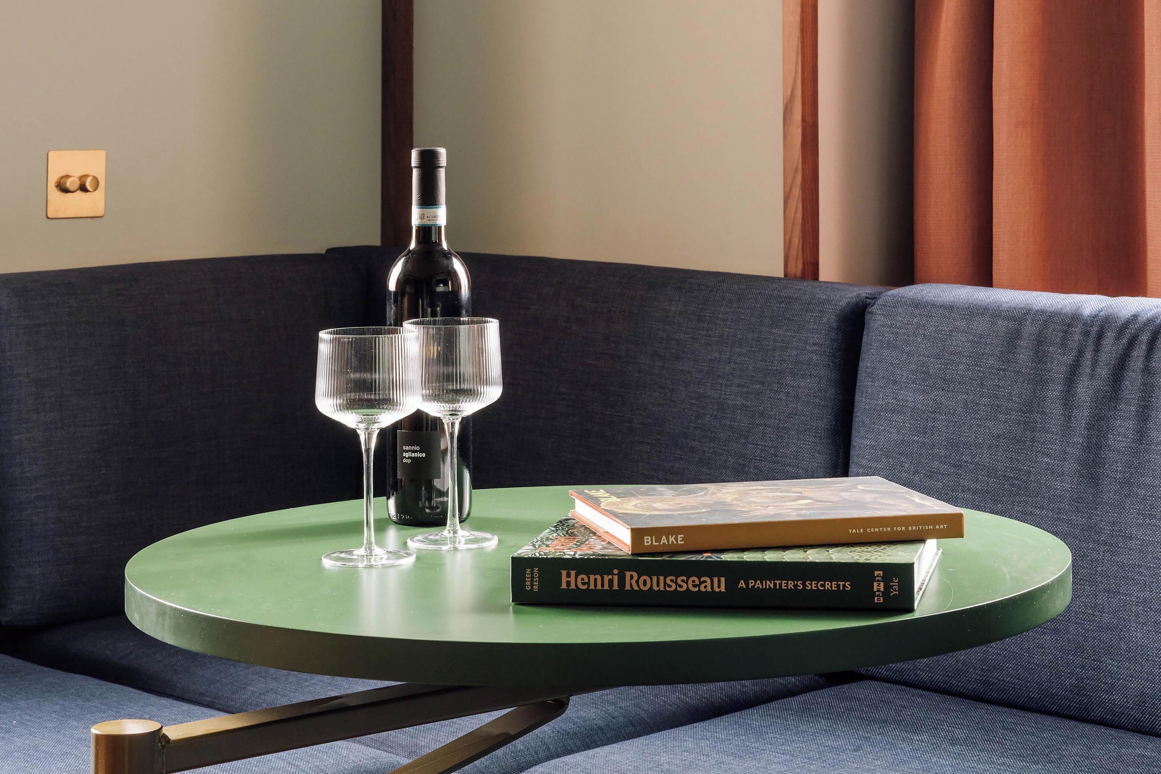 Une table ronde verte, sur laquelle sont posés deux verres à vin et une bouteille, est placée sur un canapé bleu foncé.