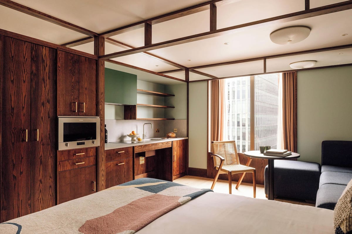Ein modernes Hotelzimmer mit einem großen Bett, einem Holzschrank, einem fest eingebauten Schreibtisch und Blick auf die Stadt durch große Fenster.