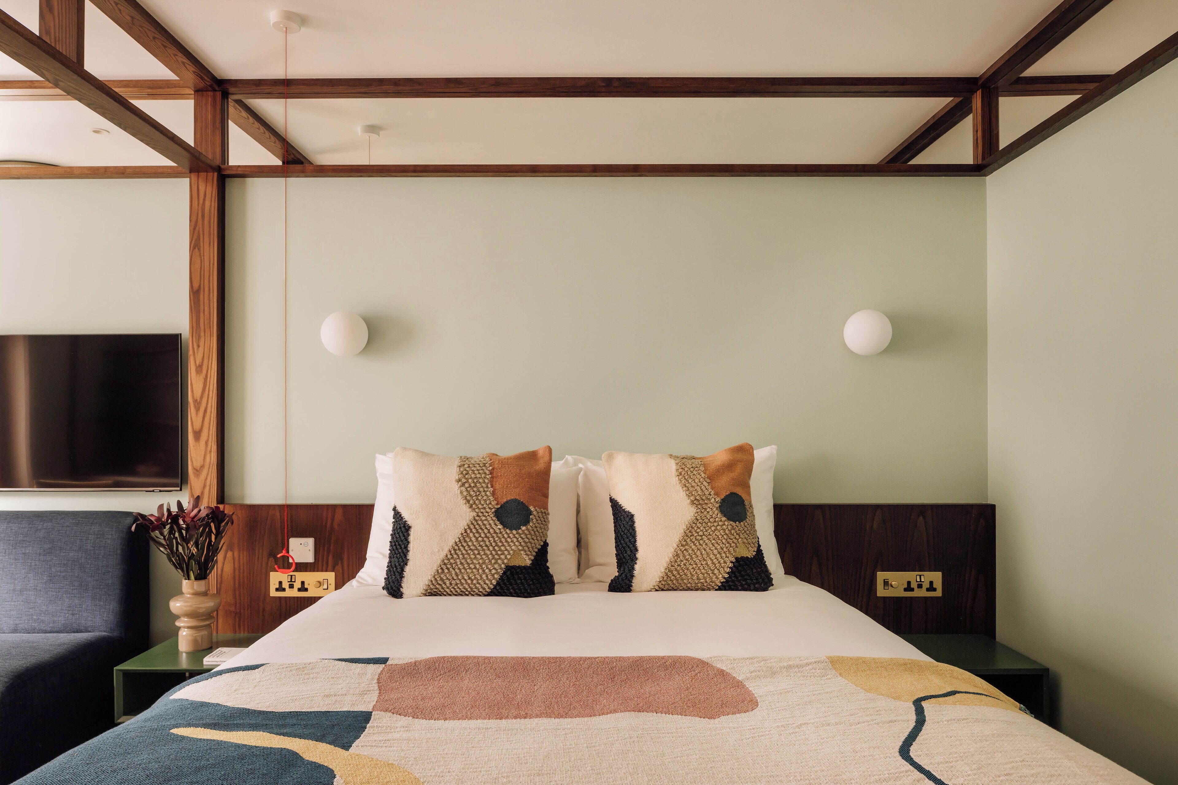 A hotel room with a bed, a nightstand, a wall-mounted TV, and a power outlet.