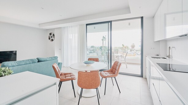 Een moderne keuken met een groot raam, een eethoek met een ronde tafel en stoelen, en een zithoek met een bank en televisie.