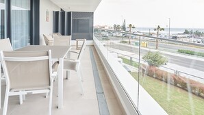 Een balkon met een tafel en stoelen, uitzicht op de straat en het strand, en een moderne gevel.