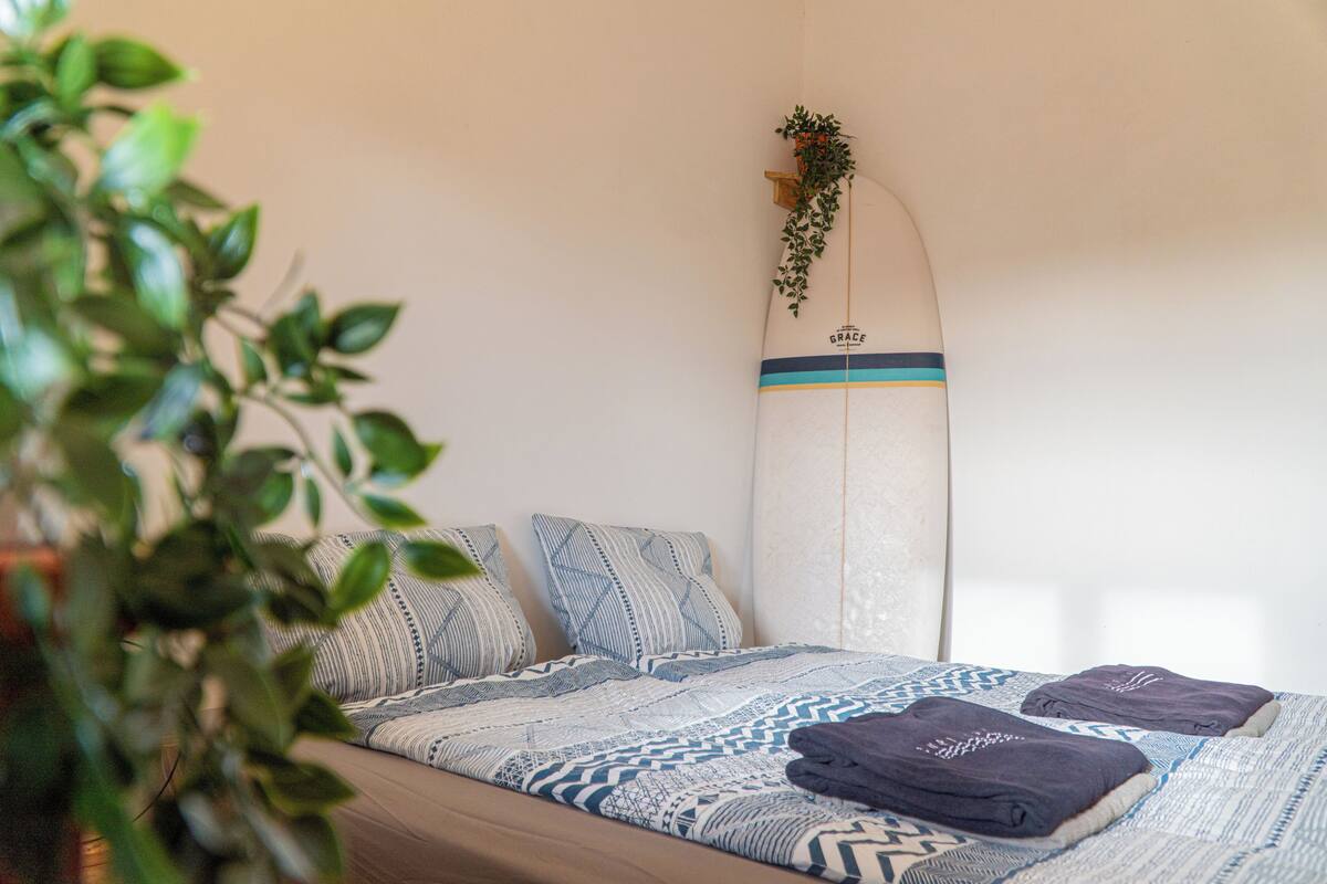 Ein Bett mit gemusterten Bettbezügen, ein Surfbrett, das an der Wand lehnt, und eine Pflanze im Vordergrund.