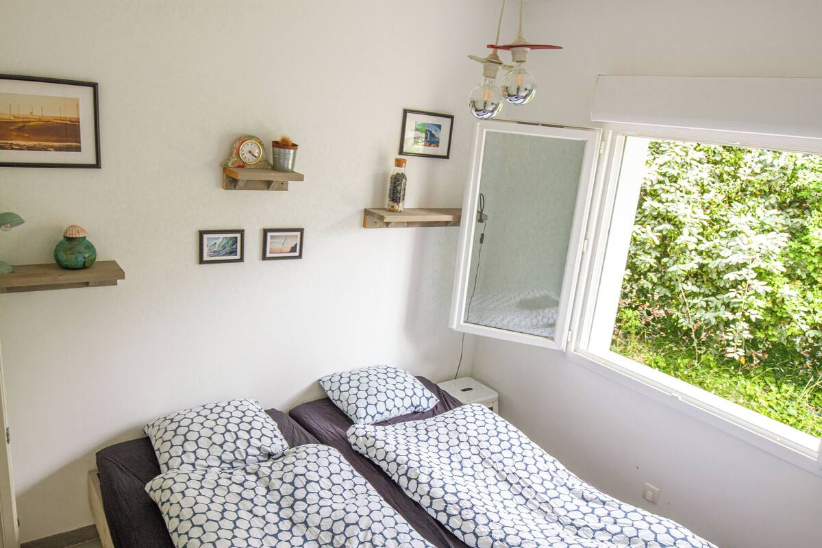 Ein kleines, gut beleuchtetes Schlafzimmer mit einem Einzelbett, einem Fenster mit Blick auf Grünflächen und mehreren an der Wand befestigten Regalen mit Dekoration.