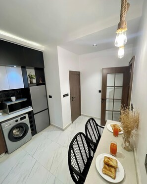A compact kitchen with modern appliances, a dining area with a table and chairs, and a hallway leading to a door. - Lakehouse Apartment (Abuja)