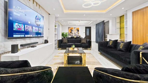 Ein modernes Wohnzimmer mit großem Flachbildfernseher, schwarzen Sofas, einem Couchtisch und einer dekorativen Deckenleuchte.