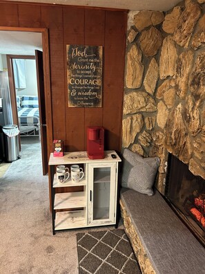 Un salon chaleureux avec un mur en pierre, une cheminée, un meuble en bois contenant des tasses et une citation inspirante sur le mur.