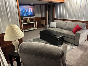A living room with a grey sofa, a black ottoman, a TV on a stand, a lamp, and a bookshelf. - Adventures Start Here-Walk to Slopes (Angel Fire)