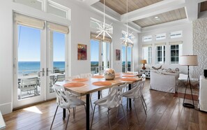 Ett rymligt matområde med ett träbord och vita stolar, stora fönster med utsikt över havet och en modern takdesign.