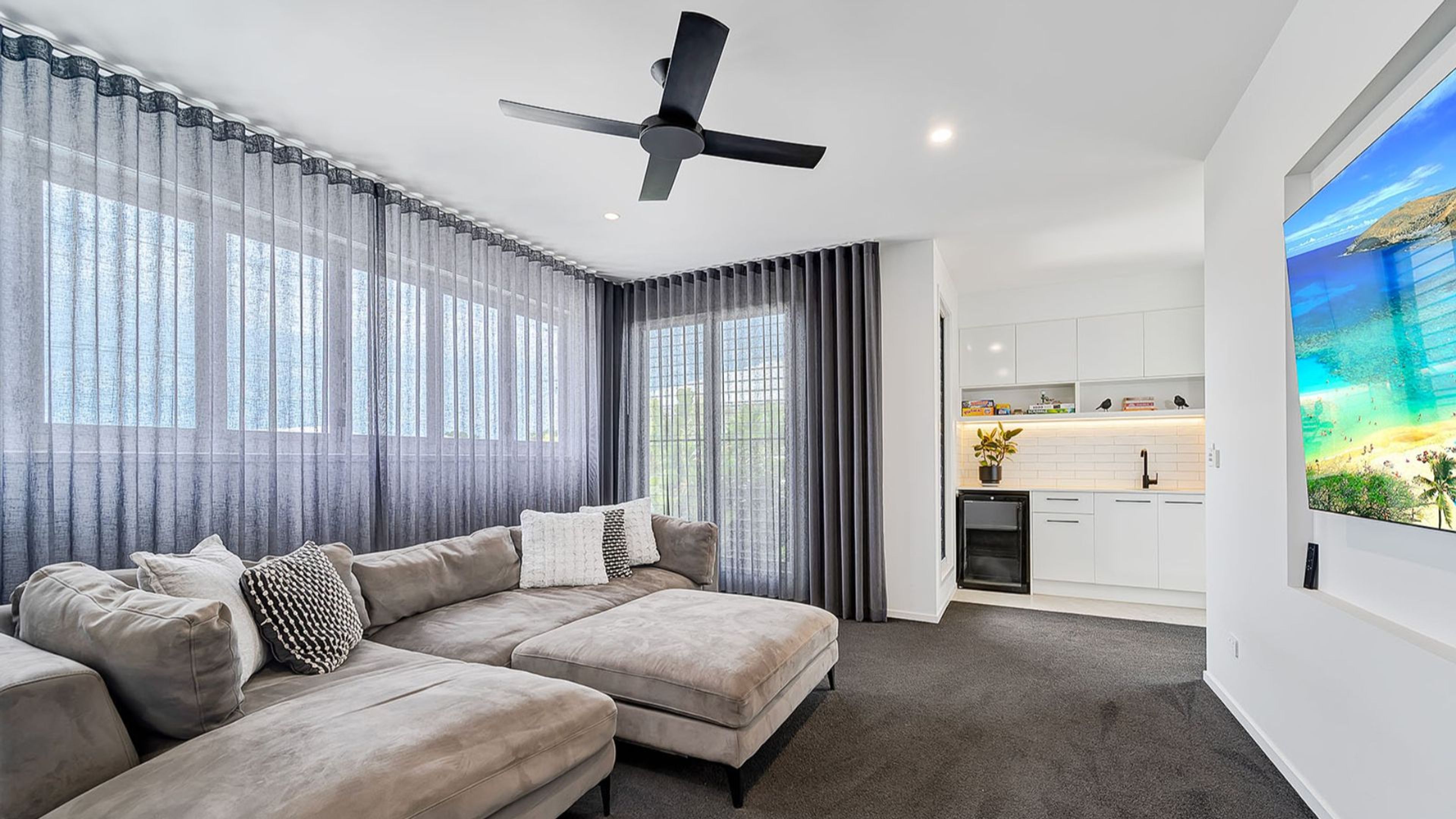 A modern living room with a sectional sofa, a ceiling fan, and a large window with sheer curtains.