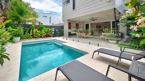 A rectangular swimming pool with a tiled deck, surrounded by lush greenery and a modern house with a glass door leading to an outdoor seating area. - Tranquil Palms Pet-Friendly Near Moffat Beach (Moffat Beach)