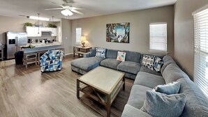 A modern living room with a sectional sofa, a coffee table, and a kitchen in the background. - Hammonds Reef | Spacious Pet-Friendly Retreat + Steps to Beach (North Padre Island)