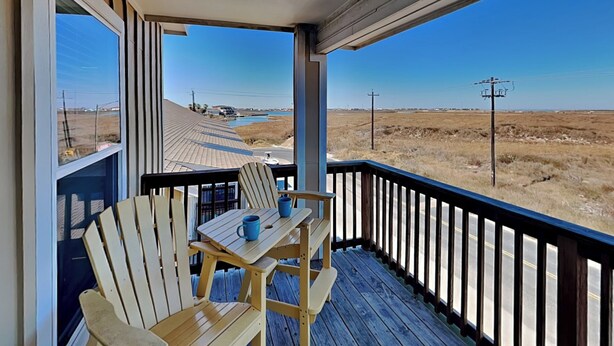 A wooden deck with two Adirondack chairs and a table, overlooking a flat landscape with a body of water and power lines in the distance. - Hammonds Reef | Spacious Pet-Friendly Retreat + Steps to Beach (North Padre Island)