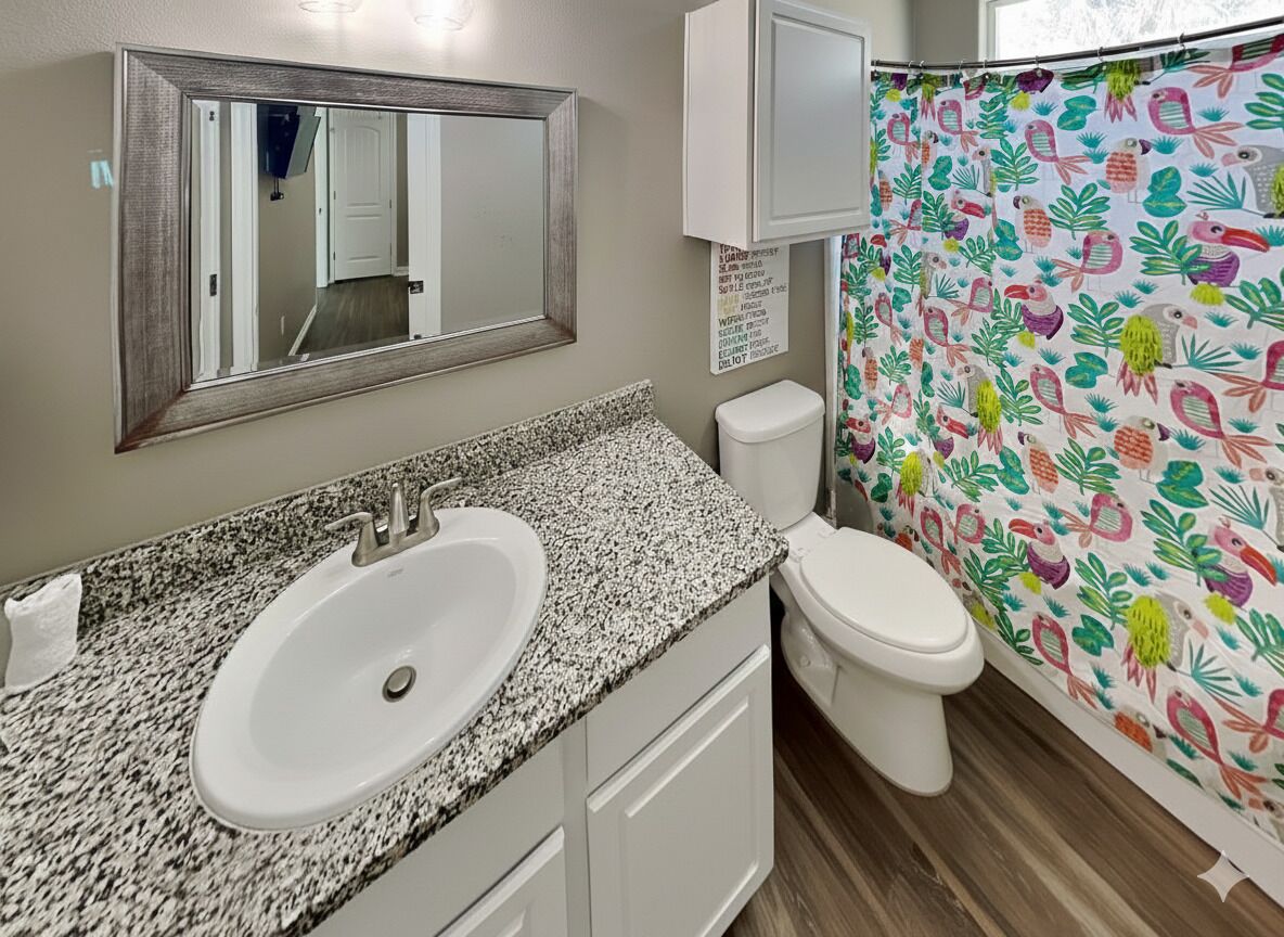 Un baño con lavamanos de encimera de granito, inodoro blanco, espejo y cortina de ducha con estampado tropical colorido.