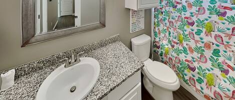 Un baño con lavamanos de encimera de granito, inodoro blanco, espejo y cortina de ducha con estampado tropical colorido.