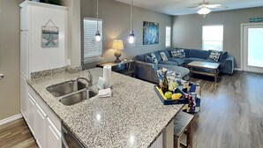 A modern kitchen with granite countertops, a built-in sink, and a dining area with a blue chair. - Hammonds Reef | Spacious Pet-Friendly Retreat + Steps to Beach (North Padre Island)