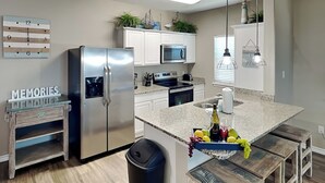 A modern kitchen with stainless steel appliances, a granite countertop, and a central island with a fruit bowl. - Hammonds Reef | Spacious Pet-Friendly Retreat + Steps to Beach (North Padre Island)