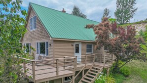 A two-story wooden house with a green roof, a deck with stairs, and a small garden area.