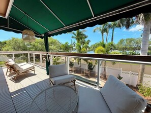 A covered outdoor deck with wicker furniture and a round table, overlooking a landscaped area with palm trees. - Luxury Hollywood Lakes Villa (Hollywood)