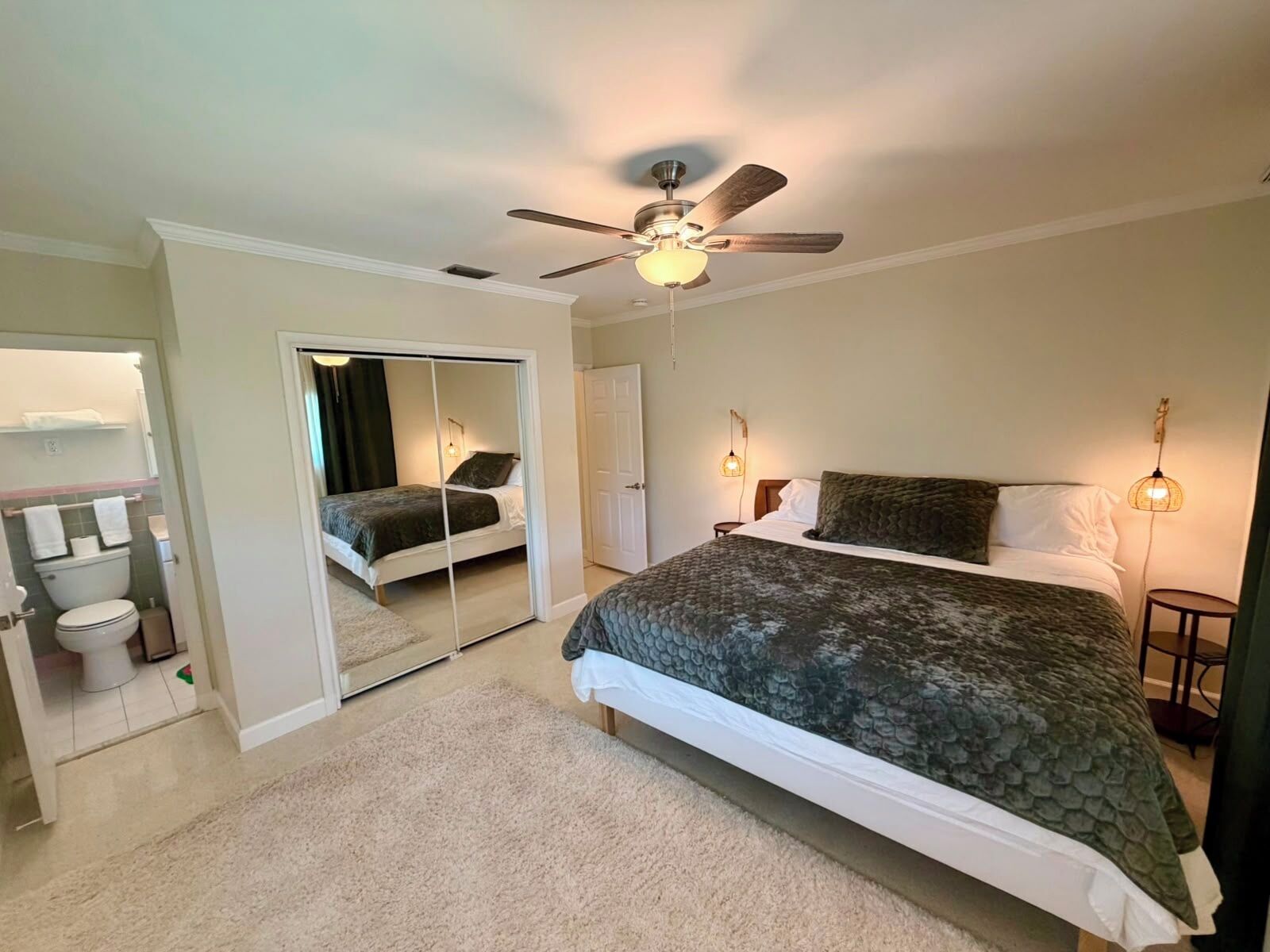 A bedroom with a bed, a ceiling fan, a nightstand, a mirror, and a bathroom with a toilet and sink.