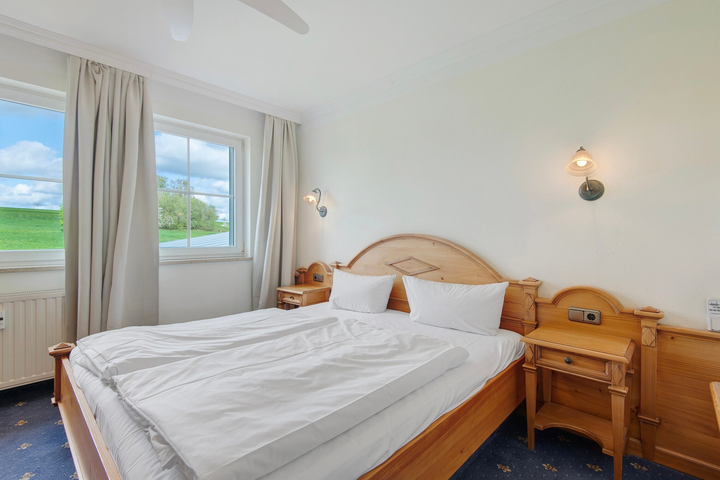 A neatly made bed with white linens, a wooden headboard, bedside tables with lamps, and a window with curtains.