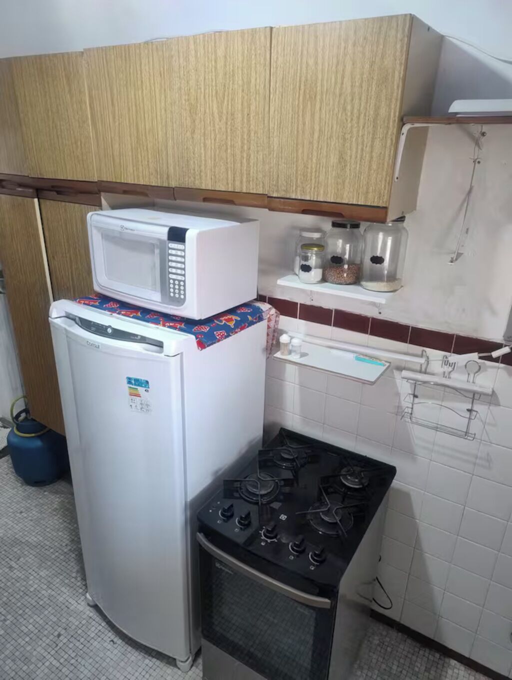 A compact kitchen with a refrigerator, microwave, gas stove, and a small shelf with jars.
