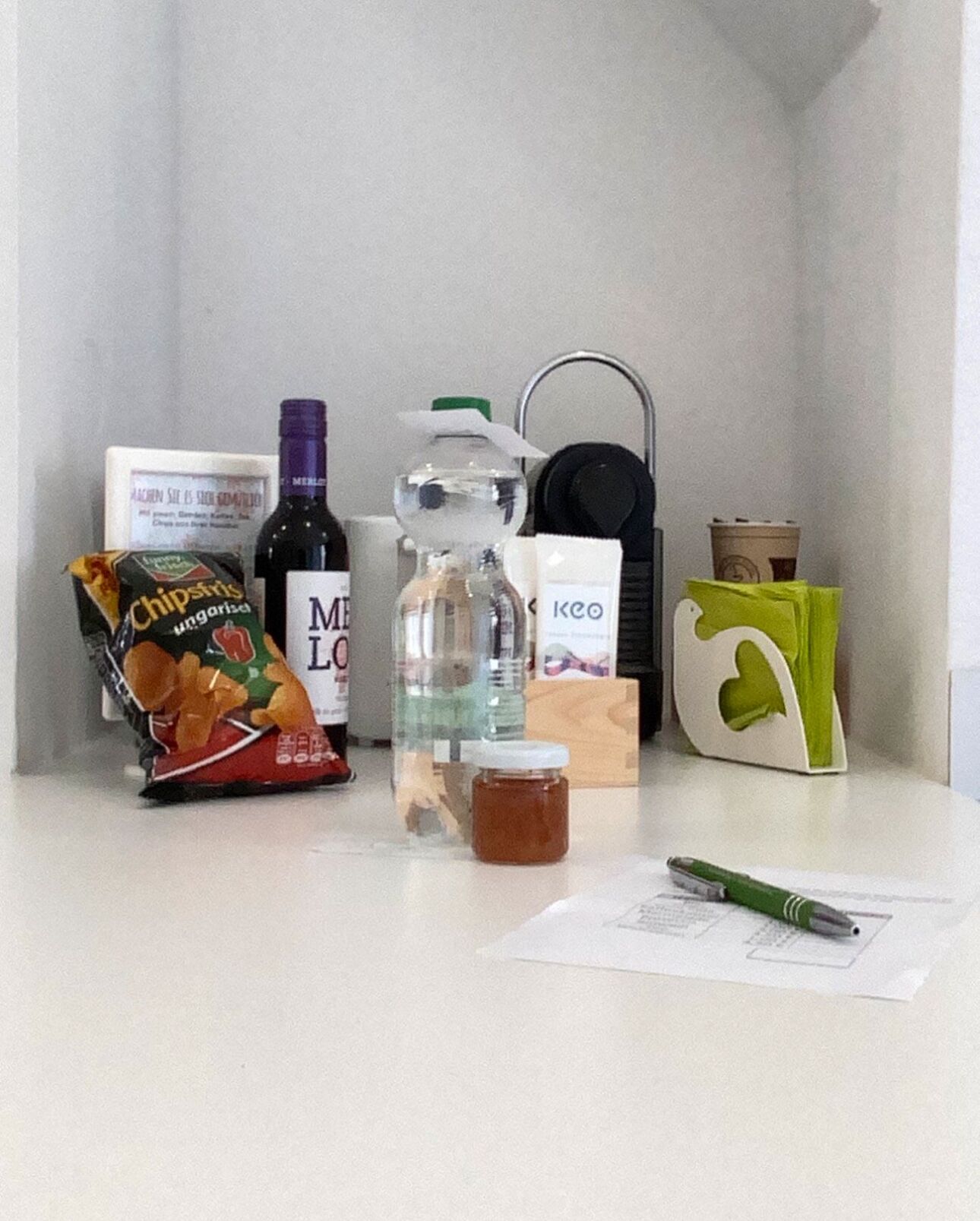 Een klein aanrecht met een zak chips, een fles Merlot wijn, een glazen pot, een groene tas en een pen op een stuk papier.