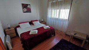 A bedroom with a bed, a bedside table, a lamp, a heater, a window with curtains, and a small table.