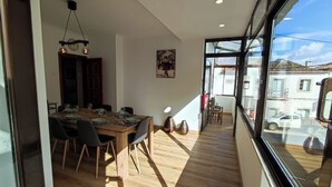A modern dining area with a wooden table and chairs, a hanging light fixture, and a large window offering a view of the street.