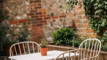 Une terrasse avec une table et des chaises, un pot de fleurs et un mur de briques orné de plantes grimpantes.