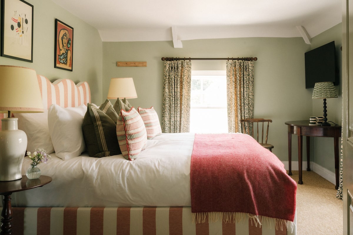 A bedroom with a bed, two bedside tables, a lamp, a vase with flowers, and a window with curtains.